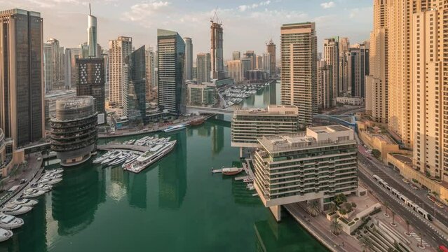 Aerial view to Dubai marina and jbr illuminated skyscrapers around canal with floating yachts night to day transition timelapse. White boats are parked in yacht club before sunrise