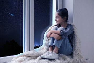 Cute little girl sitting near window and looking at shooting star in beautiful night sky