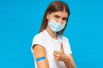Brave vaccinated woman with bandage on shoulder showing thumb up after shot on blue background - obrazy, fototapety, plakaty