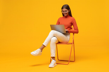 Portrait of student girl studying online sitting on chair with laptop on knees on yellow background