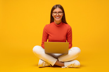 Portrait of woman on floor using laptop, playing video game, surfing internet on yellow background