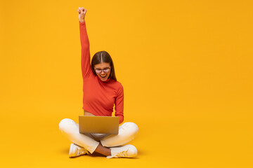 Excited business woman sitting on floor with laptop, raising hand as a winner, isolated on yellow