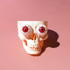 Human skull with funny eyeballs, creative arrangement against dusty rose pink background. Halloween...