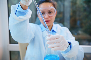Focus on laboratory glass pipette in the hands of blurred scientist, pharmacologist, clinical researcher in a white coat, safety goggles and gloves pipetting blue liquid from a flat-bottomed flask