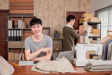 Obraz premium Young smart Asian LGBTQ men join work together partnership. Confudent male smile fashion designer sitting on desk, SME start up small business clothes store. Entrepreneur designer creative textile.