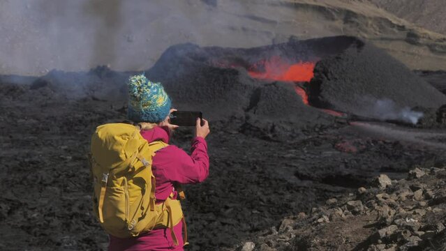 female hiker traveller on top of mountain photographing volcano erupting,adventurous excursionist woman standing on a peak taking photograph of volcanic landscape using smart phone iceland