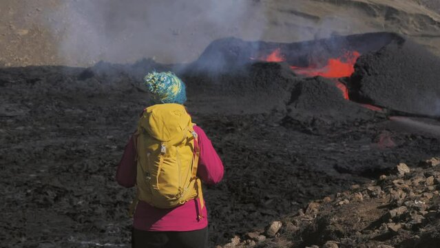 female hiker traveller on top of mountain looking at volcano erupting,travel experience,excursionist woman hiking standing on a peak looks at magma eruption volcanic landscape iceland