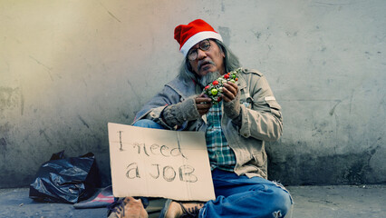 A homeless old Asian man is sitting alone by the wall laughing and drinking, holding a sign asking...