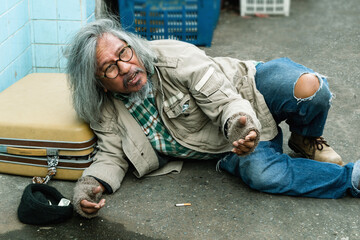 Old homeless Asian man lying on a dirty street dressed in filth reaching for help asking passersby for money or food is helpful because he has no work and no home.