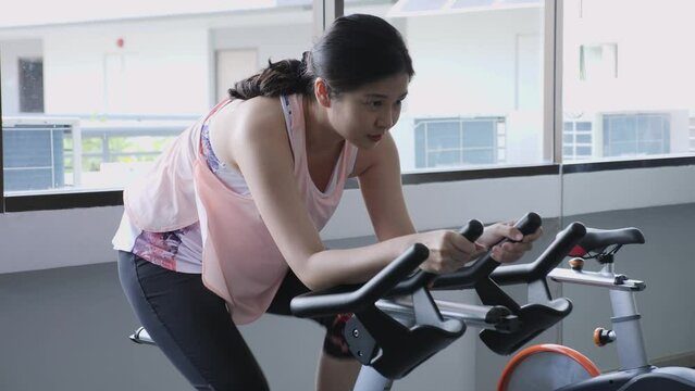 Medium shot of middle-aged Asian woman, exercising, sprinting, using indoor cycling machine, workout alone at indoor fitness gym. Sport, health, wellbeing, weight loss concept.
