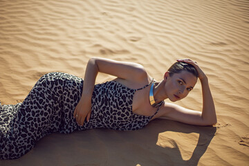 brunette woman in a long leopard dress lie with her back in the desert at sunset. go everywhere.