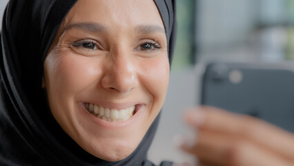 Headshot young pretty happy arab woman in hijab smiling with white healthy teeth looking at smartphone screen rejoicing at good news watching funny video on telephone receives positive result by email