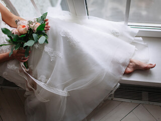 The bride is sitting on the windowsill by the window in a white lace dress with embroidery, holding...