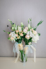 Two bouquets in light colors of white eustoma, white and cream roses, wrapped in blue ribbons, stand in a glass vase.