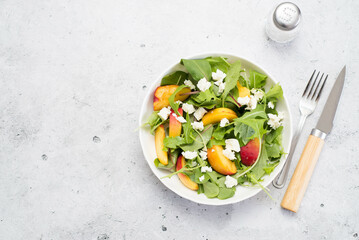 A light summer salad of rucolla, peaches and cheese spoon and knife on a light gray background.