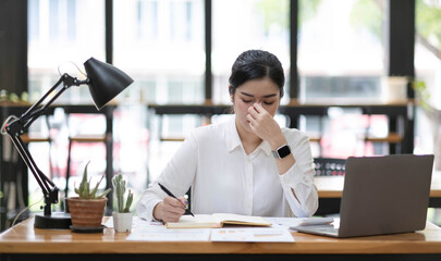 Young Asian woman feeling migraine head strain. Tired, Overworked businesswoman financier while working on laptop computer in office.