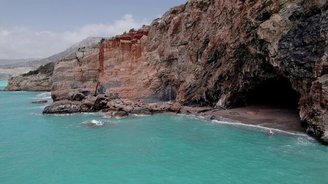 Aerial view of family supboarding to cave at secluded rocky beach. Mom and dad oaring on paddleboards with their sons down azure sea