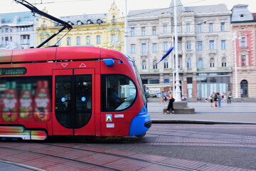 closeup of red and blue tram in the popular Jelacic square in downtown Zagreb. old architectural background. tourism and travel to croatia concept. soft blurred urban background with tourists - obrazy, fototapety, plakaty