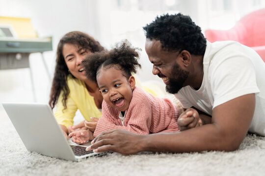 African American family laying down on the floor and looking at laptop with daughter with smile in the living room. Happy family day on vacation. Kid feeling happy and fun during playing game.