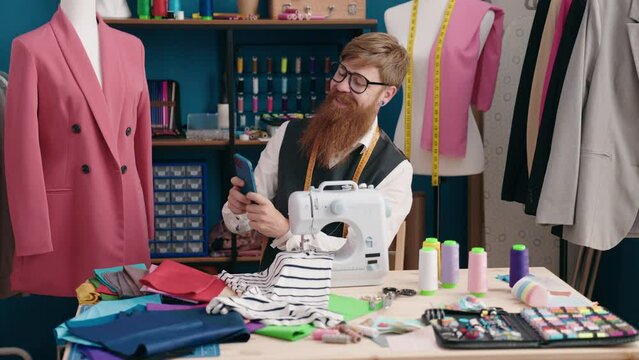 Young redhead man tailor using sewing machine make selfie by smartphone at clothing factory