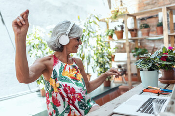 Cheerful senior woman listening to music and using her laptop while sitting in greenhouse.