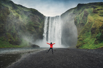 Male tourist in red jacket enjoying the Skogafoss waterfall flowing from cliff on summer in Iceland