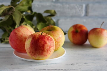 big red apples are lying on a white plate on a wooden table with the fruits of scattered apples and a branch with green leaves on the background of a brick white wall