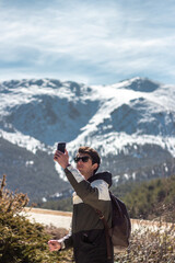 Handsome caucasian man posing to camera. He carries his cell phone and sunglasses. he has snowy mountains in the background