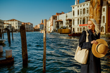 Fototapeta na wymiar Elegant happy smiling traveler woman wearing blue trench coat, holding straw hat, with big hobo bag, posing in street of Venice. Fashion, travel, lifestyle conception. Copy, empty space for text