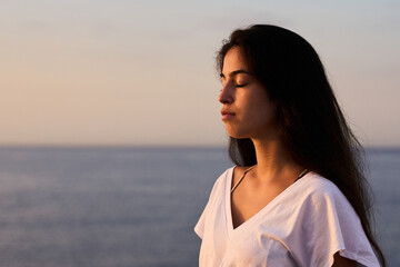 Obraz premium young hispanic tanned girl with a white t-shirt meditating at the rocks early in the morning in the seaside