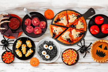 Fun Halloween dinner party table scene over a white wood background. Overhead view. Ghost pizza, eyeball spaghetti, jack o lantern pumpkin pie, candy apples, snacks and spooky punch.