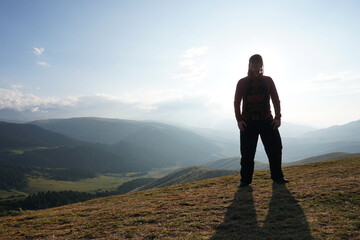 A guy on the edge of the hill admires the view. In the distance you can see high mountains and green hills covered with forest. Wide margins. Clouds are visible and sun is shining brightly