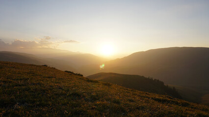 Sunset sun over yellow-green hills. Small clouds are illuminated by the orange-red rays of the outgoing sun. A light haze in the gorge. Coniferous trees grow. Clear sky. Assi-Turgen Plateau Kazakhstan