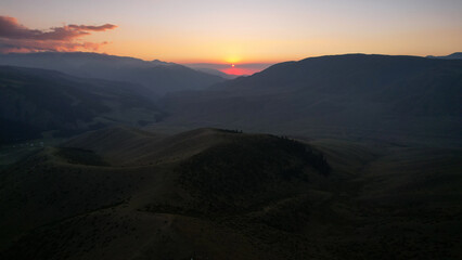 Red rays of the sun at sunset among the mountains. Green grass turns yellow, in places a small forest. Tourists walk in the hills. A river runs in the distance. The clouds are highlighted in pink.