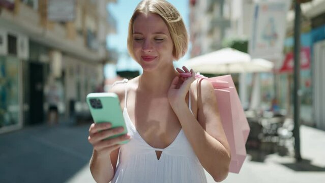 Young blonde woman using smartphone holding shopping bags walking at street