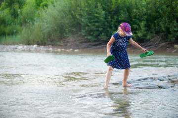 The girl stands by a small clear mountain river and tries to cross it.