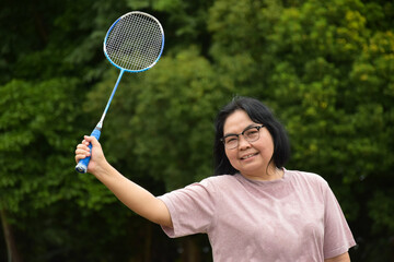 Portrain of asian middle aged woman holding badminton racket, standing and smiling in the green field to play outdoor badminton, concept for to be strong and healthy woman by exercising afterwork.