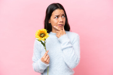 Young Colombian woman holding sunflower isolated on pink background having doubts and thinking
