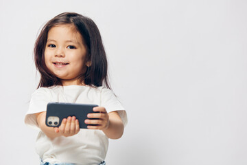 cute, funny, little girl in a white t-shirt stands on a light background with a smartphone in her hand and takes a selfie. Horizontal studio photography with blank space for advertising mockup insert