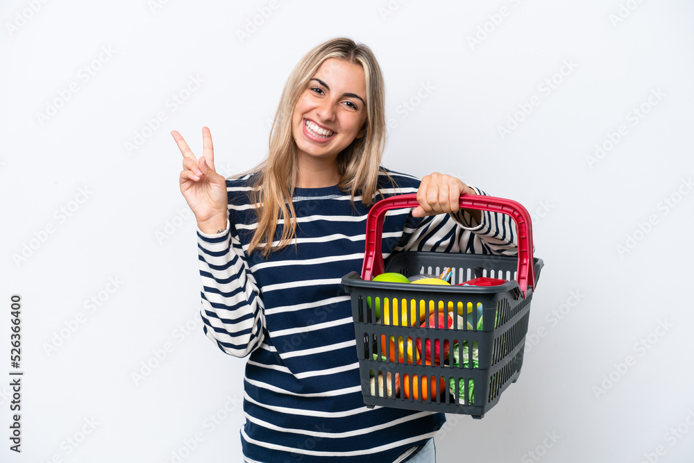 Wall mural Young caucasian woman holding a shopping basket full of food isolated on white background smiling and showing victory sign