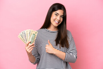 Young Brazilian woman taking a lot of money isolated on pink background giving a thumbs up gesture