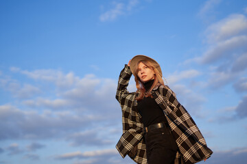 Attractive blonde long hair woman in stylish clothes - shirt and brown hat on with blue cloudy sky background. Portrait of a girl looking forward. High quality image