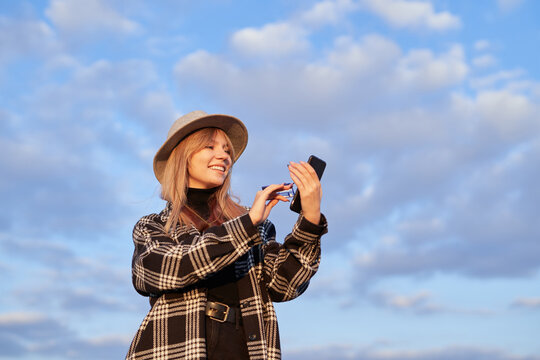 Attractive hipster girl making online shopping in background of blue cloudy sky. Young caucasian blonde female in hat holding smartphone and credit card. High quality image