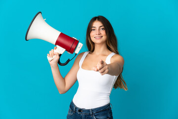 Young caucasian woman isolated on blue background holding a megaphone and smiling while pointing to the front