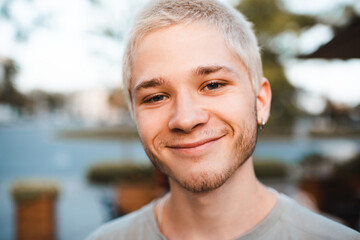 Handsome young man with blond short cut hairstyle smiling over city urban background outdoor. Look at camera. Close up portrait of 18-19 year old happy teenage boy.