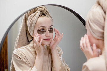 A well-groomed woman at home in a bathrobe and towel applies patches under the eyes from swelling...