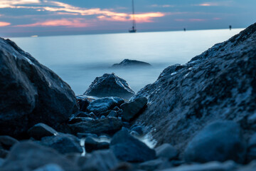 Sonnenuntergang am Meer mit Segelbooten im Hintergrund und Steinen im Vordergrund mit Langzeitbelichtung