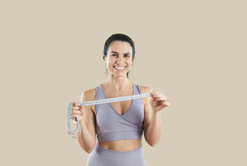 Happy female sports trainer or fitness instructor holding measuring tape isolated on beige background. Portrait of caucasian middle-aged athletic woman with beautiful smile dressed in sportswear.