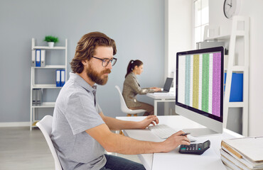 Male accountant uses calculator to calculate financial report working at office with computer. Man working with financial software sitting at desk on colleague background. Finances and economy concept