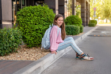 A beautiful teenage girl with a backpack is sitting on the road  before school. The concept of returning to school, education and childhood